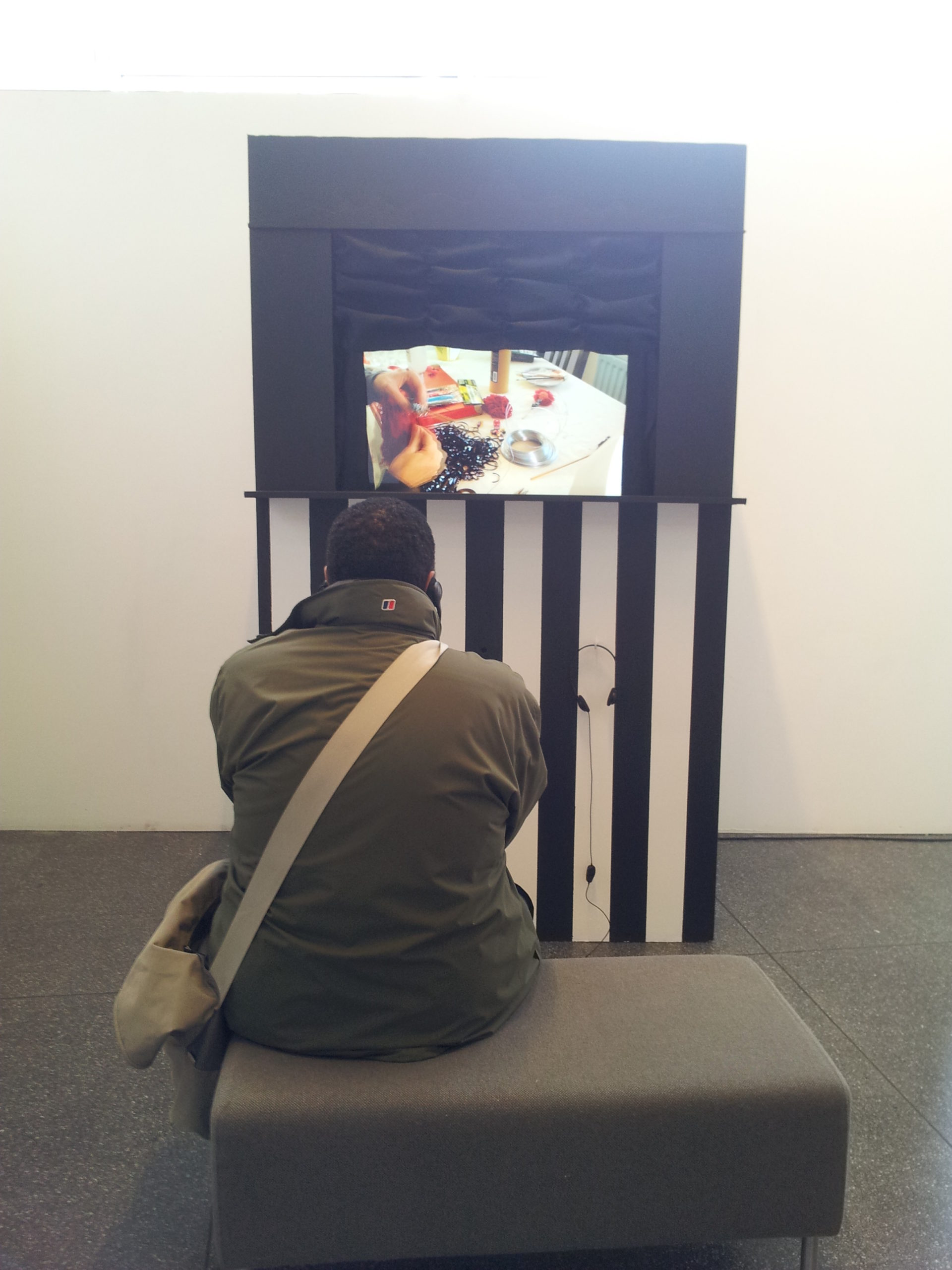 a man sits on a bench watching a screen set into a booth that is decorated with black and white stripes