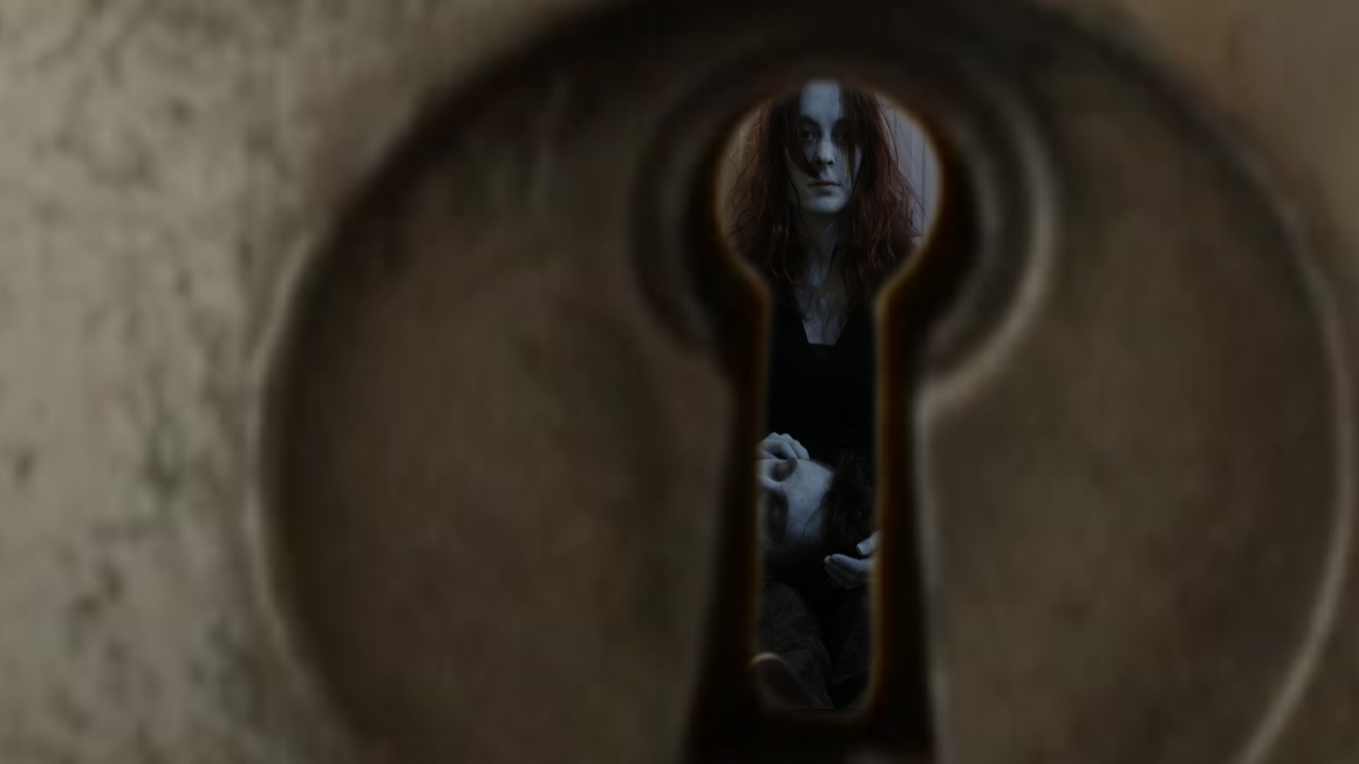 a keyhole with an image through the keyhole of a zombie-like woman sitting with back against the wall and a zombie-like man with his head in her lap