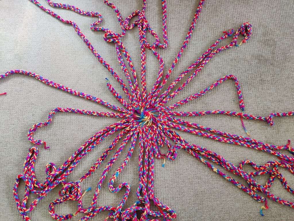 Image shows strands of finger knitted wool of differing lengths arranged in a circle with the ends at the centre in a spiral. The wool is multi-coloured.