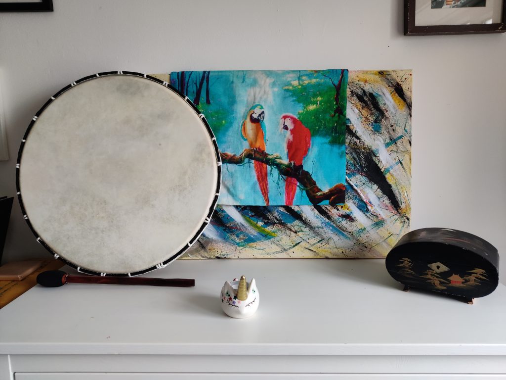picture shows a painting with a cushion cover with an image of 2 parrots on it draped over the painting, with a shamanic drum resting against the painting, a black jewellery box, a drum beater and a candle holder in the shape of a cat face with a unicorn horn