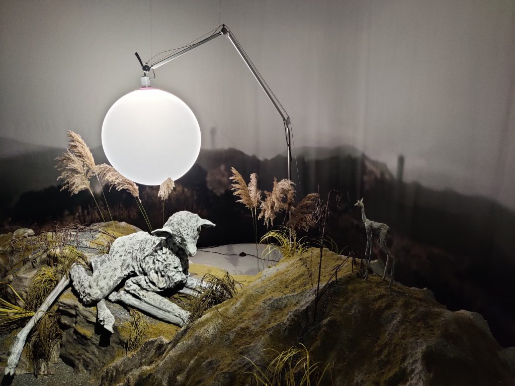 Image shows an art exhibit with sculpted landscape with a sheep with elongated legs laying down with legs folded up, a tiny deer (in relation to the sheep's - a tenth of the size) and a spherical light hanging above.