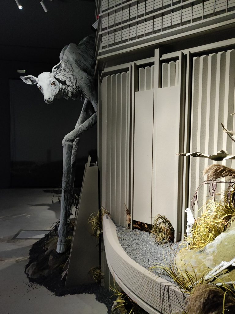 Image shows an art exhibit with a sheep standing on elongated legs (like stilts) peering around the corner of a metal structure in a dimly room.