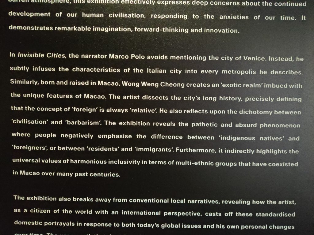 Image shows a photo of text taken from the Above Zobeide art exhibition that reads: “barren atmosphere, this exhibition effectively expresses deep concerns about the continued development of our human civilisation, responding to the anxieties of our time. It demonstrates remarkable imagination, forward-thinking and innovation.

In Invisible Cities, the narrator Marco Polo avoids mentioning the city of Venice. Instead, he subtly infuses the characteristics of the Italian city into every metropolis he describes. Similarly, born and raised in Macao, Wong Wong Cheong creates an ‘exotic realm’ imbued with the unique features of Macao. The artist dissects the city’s long history, precisely defining that the concept of ‘foreign’ is always ‘relative’. He also reflects upon the dichotomy between ‘civilisation’ and ‘barbarism’. The exhibition reveals the pathetic and absurd phenomenon where people negatively emphasise the difference between ‘indigenous natives’ and ‘foreigners’, or between ‘residents’ and ‘immigrants’. Furthermore, it indirectly highlights the universal values of harmonious inclusivity in terms of multi-ethnic groups that have coexisted in Macao over many past centuries.

The exhibition also breaks away from conventional narratives, revealing how the artist, as a citizen of the world with an international perspective, casts off these standardised domestic portrayals in response to both today’s global issues and his own personal changes”