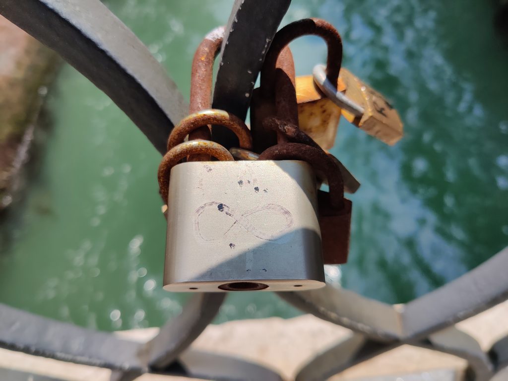 Image shows a close up shot of a padlock chained to a the metalwork of a bridge. The padlock has a figure of 8 symbol drawn onto it.