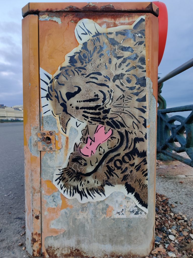 A jaguar or leopard wheatpasted onto an orange metal storage cupboard, rusty in places, on Hove esplanade, with the fence and the beach beyond to the right and to the left, apartment buildings overlooking the seafront
