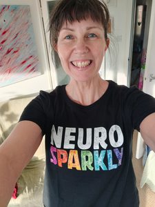 Julia, a white woman with blue eyes and brown fringe is wearing a black t shirt with the words "neuro sparkly" on the front, and is beaming at the camera.