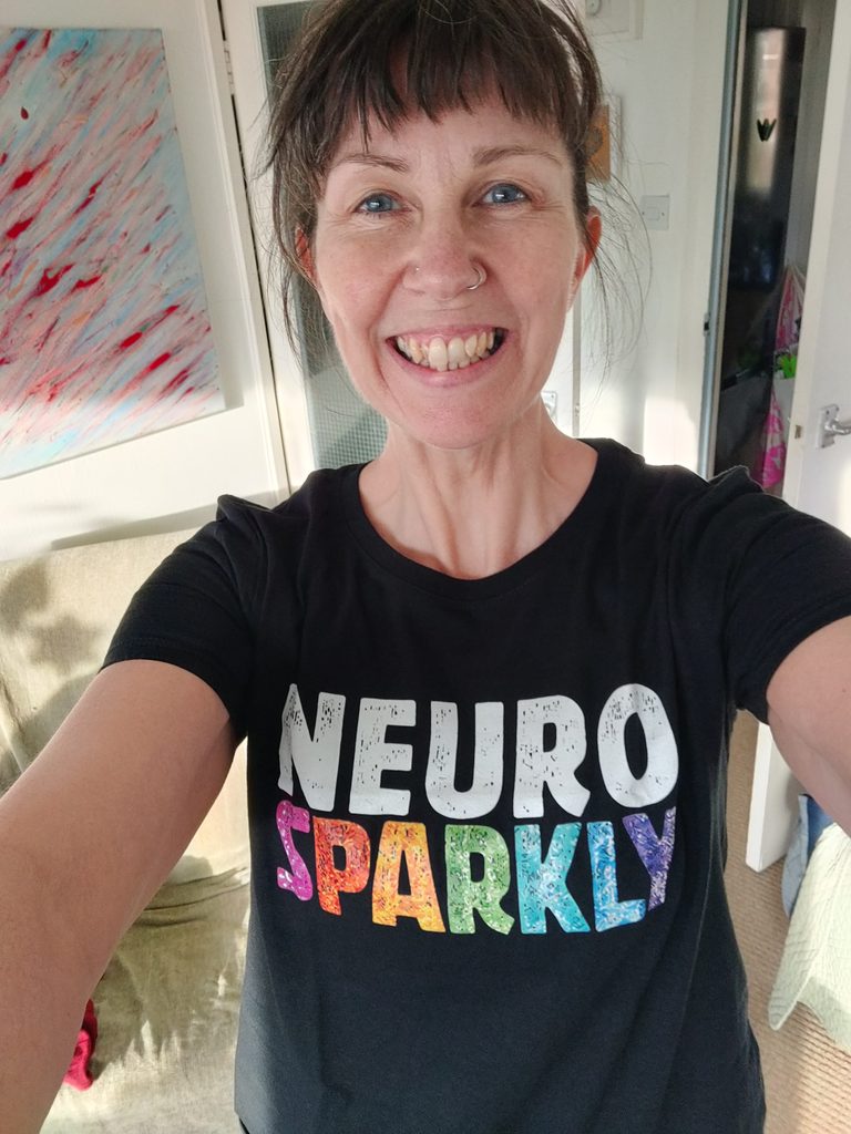 Julia, a white woman with blue eyes and brown fringe is wearing a black t shirt with the words "neuro sparkly" on the front, and is beaming at the camera.